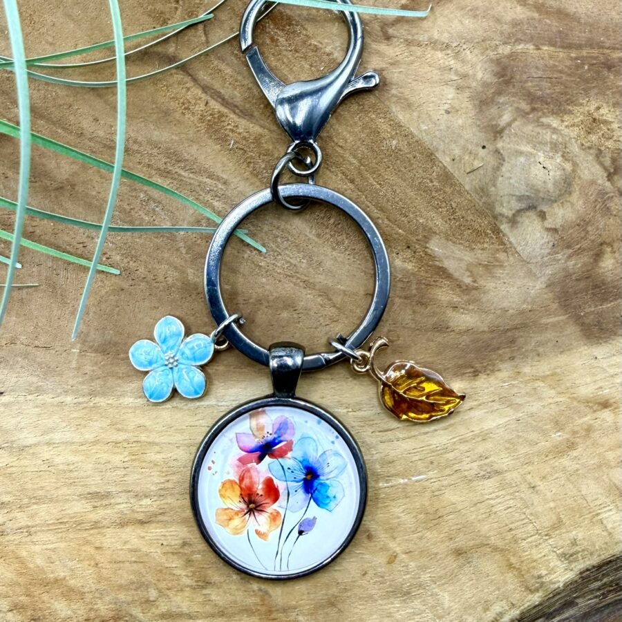 Craquez pour ce porte-clés fleurs en acier inoxydable avec cabochon de 25 mm et deux breloques assorties. Une création artisanale colorée et originale.